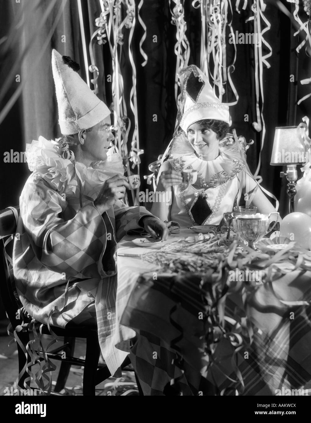 1920s 1930s SMILING COUPLE DRINKING COFFEE FROM CHINA CUPS WEARING HARLEQUIN COSTUMES AMID PARTY STREAMERS Stock Photo