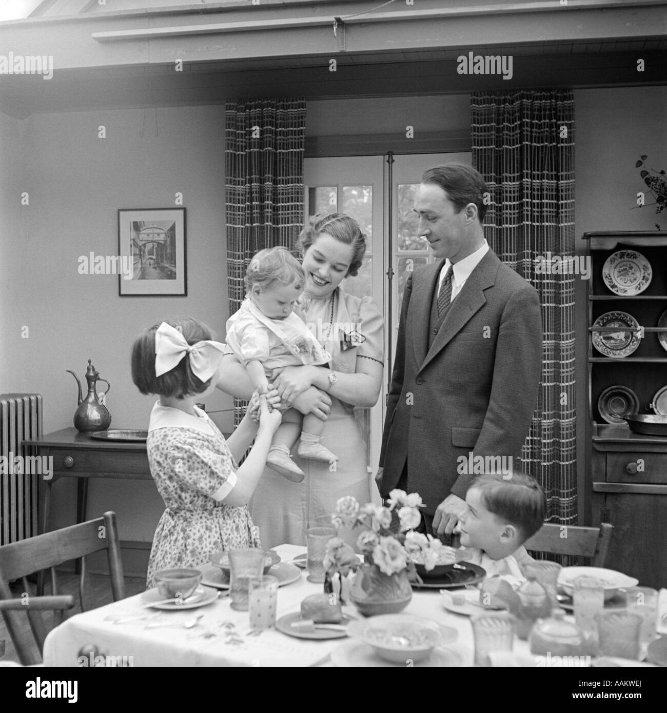 1930s FAMILY OF 5 IN DINING ROOM MOTHER STANDING HOLDING BABY Stock Photo