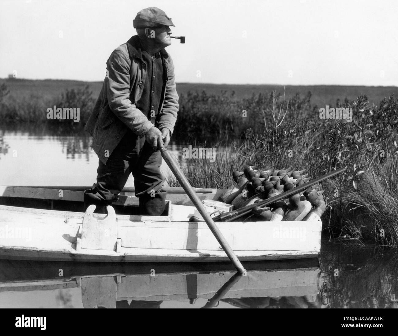 1920s 1930s SENIOR MAN DUCK HUNTER STANDING IN SNEAK-BOX BOAT ABOUT TO DEPLOY DECOYS IN  BARNEGAT BAY WETLANDS MARSH NEW JERSEY Stock Photo