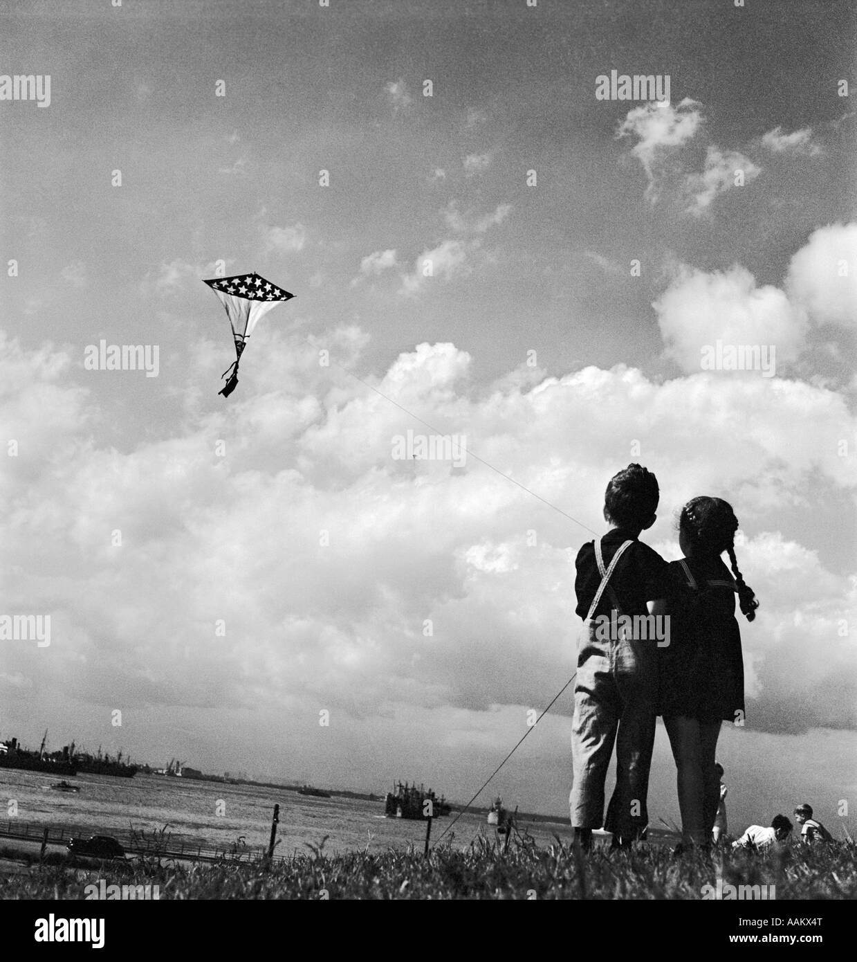 1940s YOUNG GIRL AND BOY STANDING SIDE BY SIDE FLYING PATRIOTIC FLAG KITE OVER RIVER Stock Photo