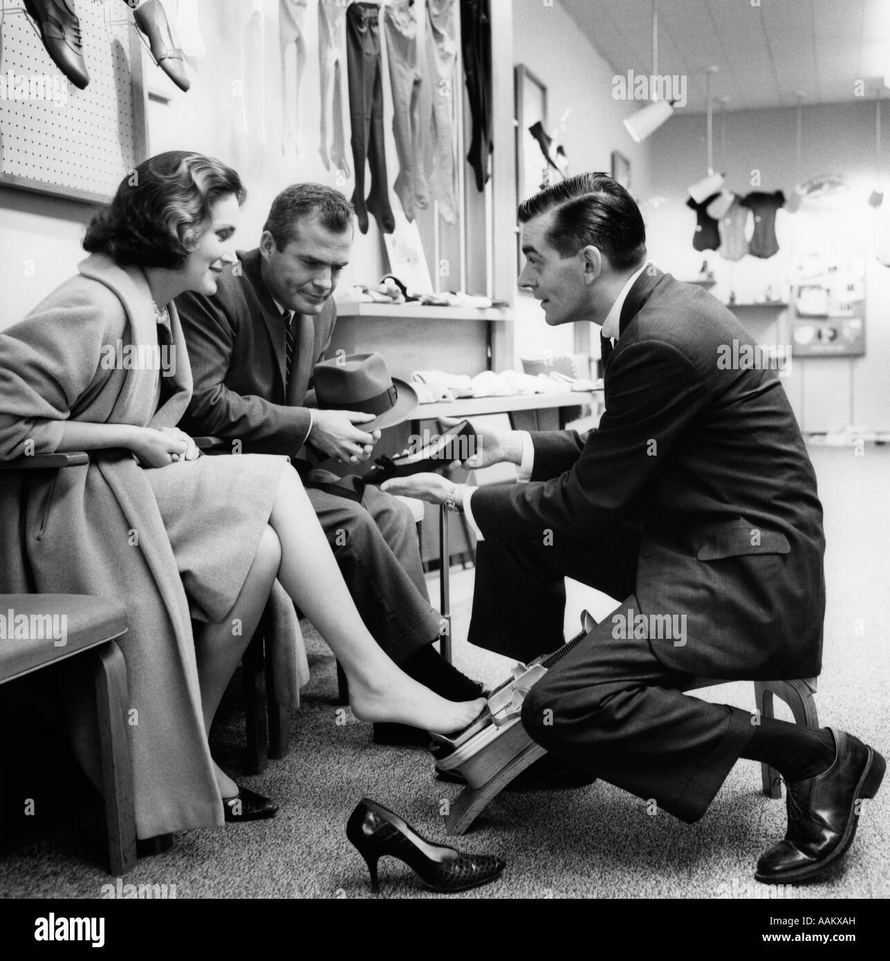 1950s 1960s COUPLE WOMAN TRYING ON SHOES MAN LOOKING IMPATIENT SHOE SALESMAN DOING HIS JOB Stock Photo