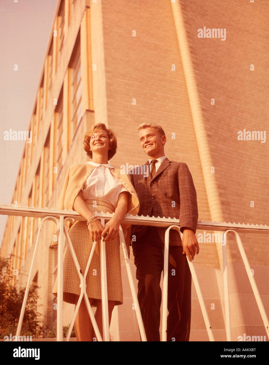 1950s 1960s YOUNG SMILING BLONDE TEENAGE COUPLE STANDING TOGETHER OUTDOORS BY WROUGHT IRON RAILING AND BRICK BUILDING Stock Photo