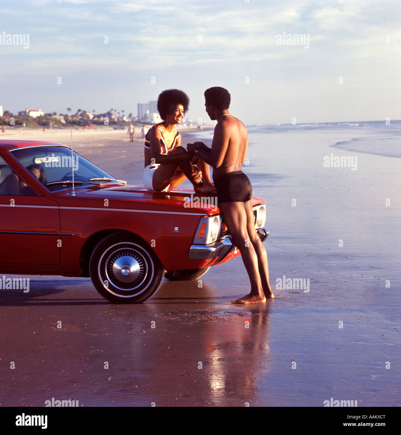 1970s SMILING ROMANTIC AFRICAN AMERICAN VACATION COUPLE AT BEACH WOMAN SITTING ON CAR HOOD MAN HOLDING HANDS Stock Photo