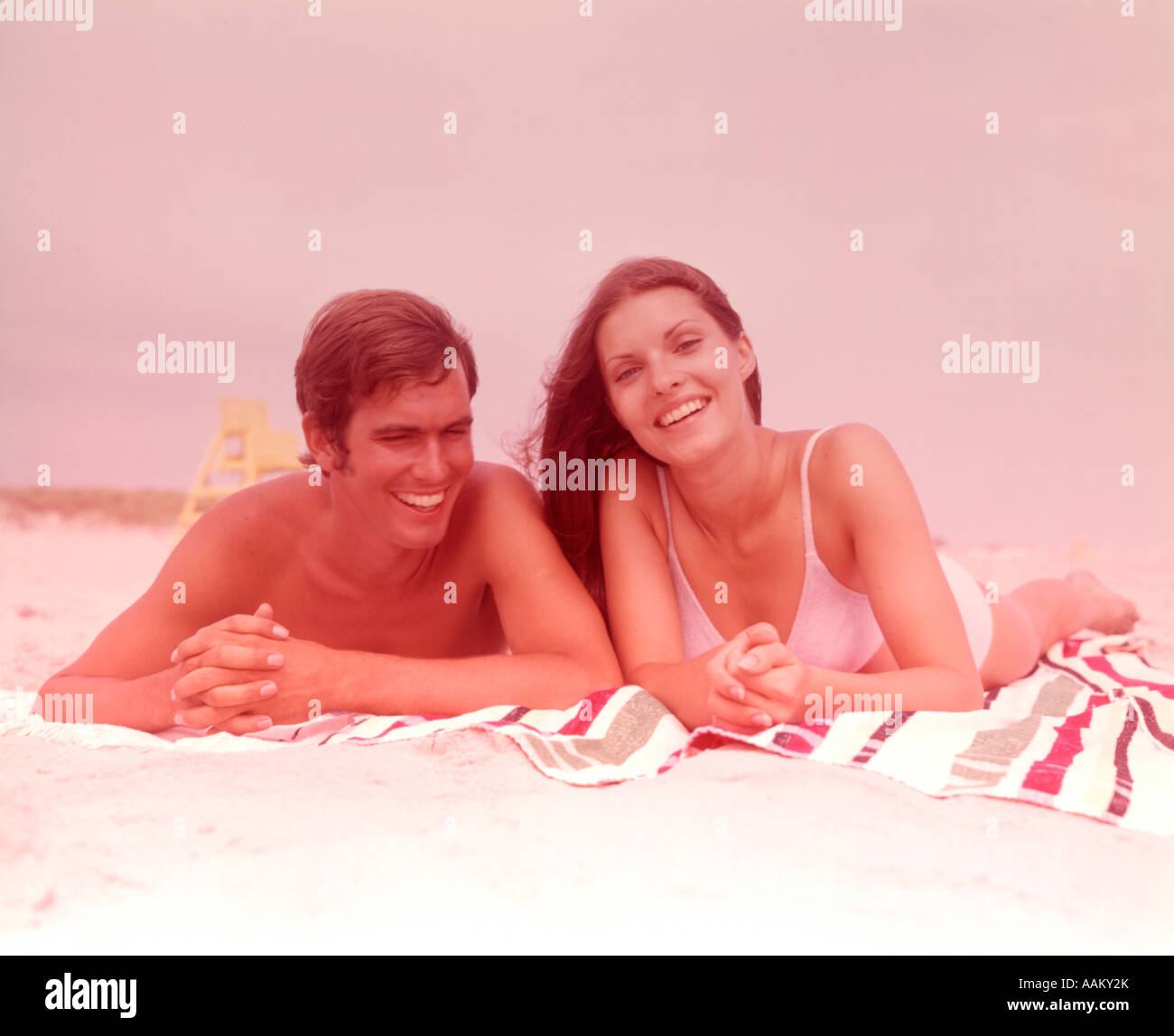 1960s SMILING YOUNG COUPLE LYING SIDE BY SIDE TOGETHER ON BEACH TOWEL Stock Photo