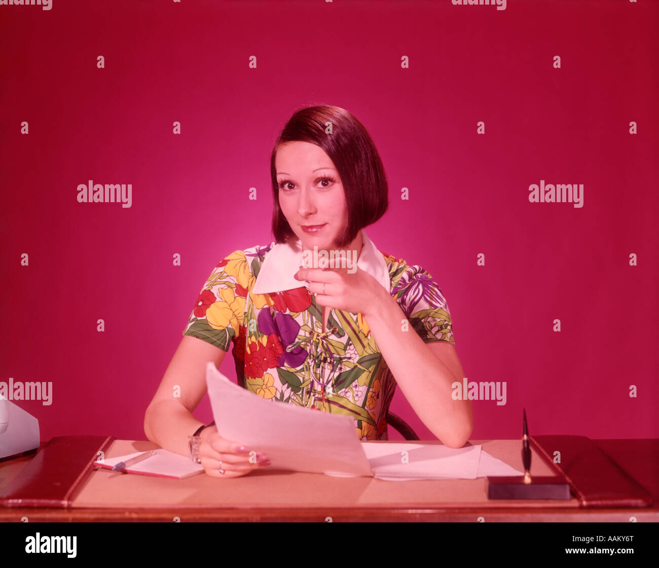 1970s WOMAN AT DESK COLORFUL BLOUSE POINTING TOWARDS LOOKING AT CAMERA Stock Photo