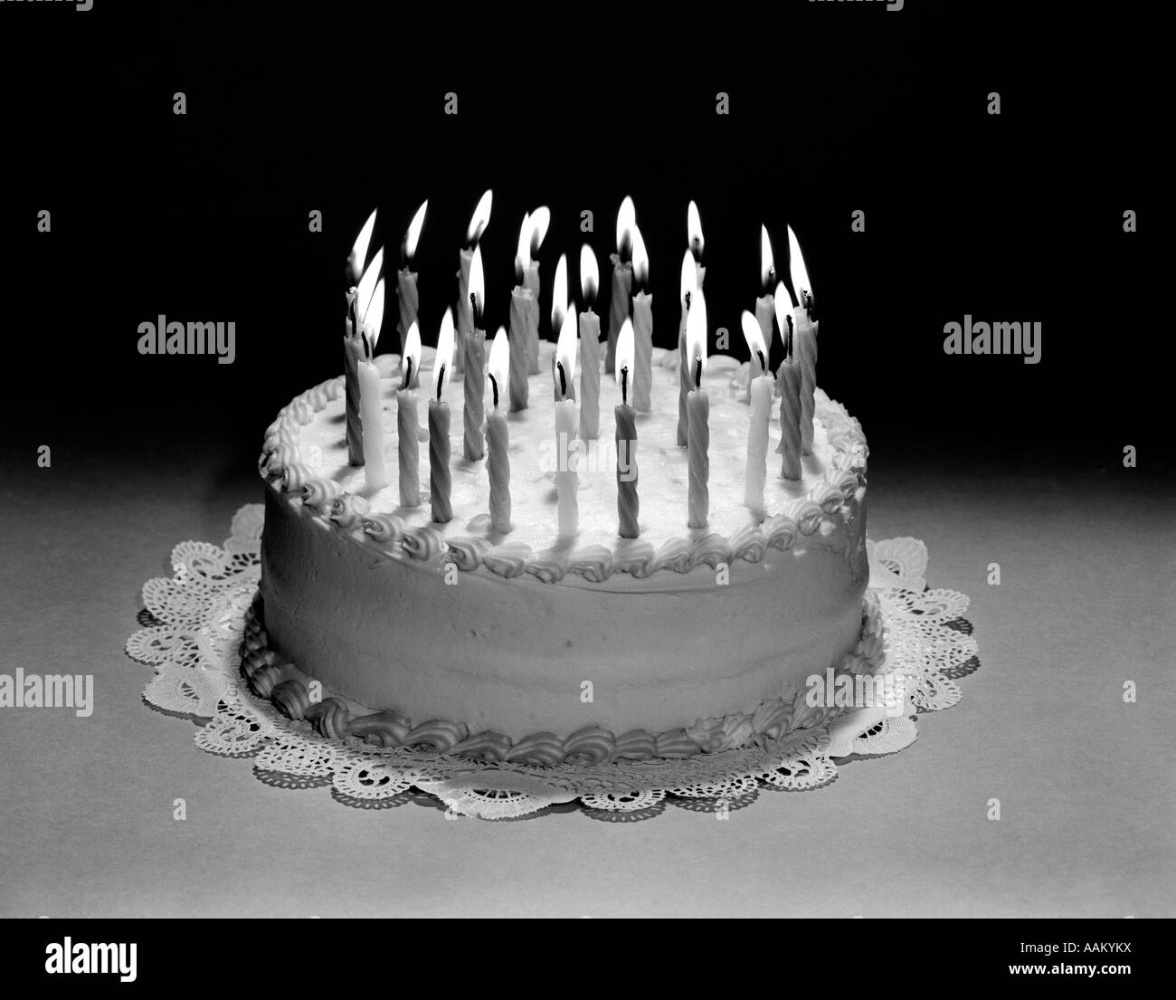 1960s BIRTHDAY CAKE STILL LIFE WITH MANY LIT CANDLES ON TOP OF PAPER DOILY Stock Photo