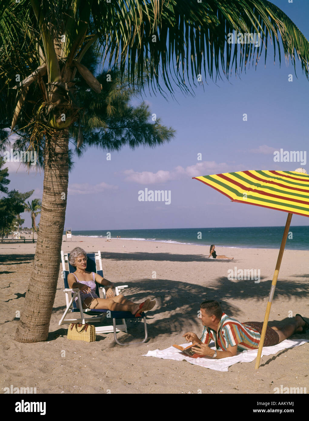 1960s MAN AND WOMAN COUPLE SUNBATHING ON BEACH IN FLORIDA TALKING TOGETHER UNDER PALM TREE AND COLORFUL BEACH UMBRELLA Stock Photo