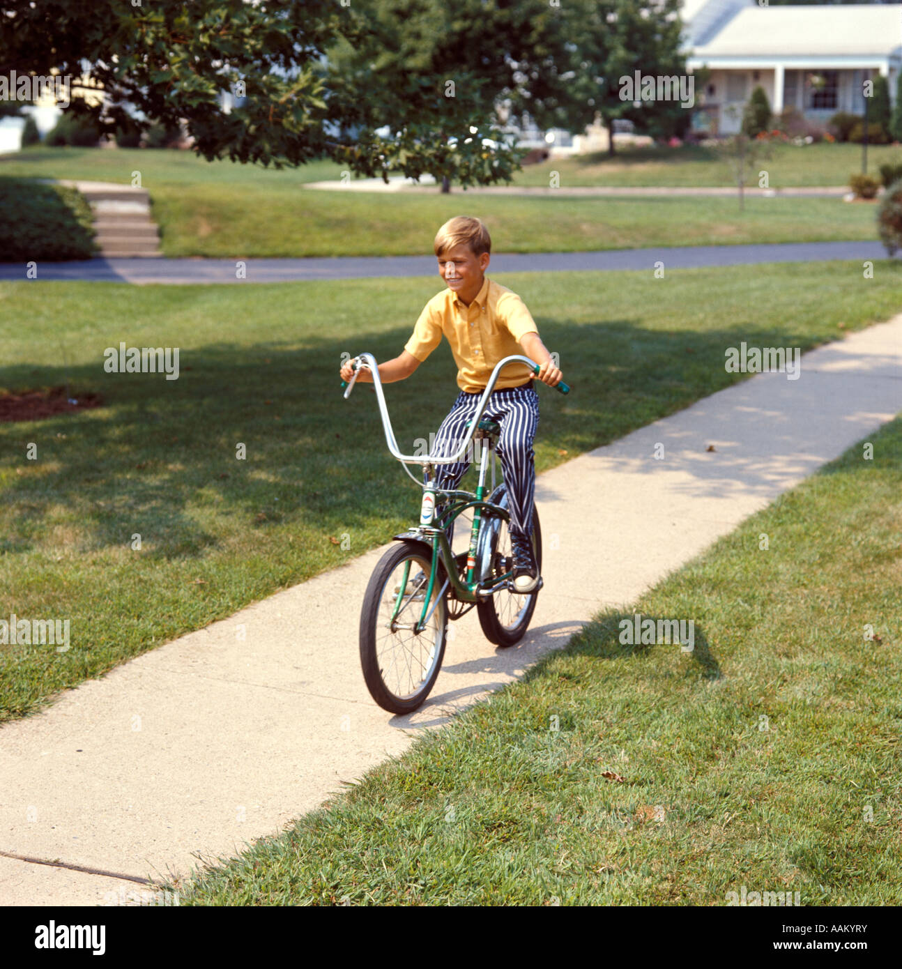 1970s BLOND BOY BIKING WITH CHOPPER HANDLEBARS RIDING DOWN SIDEWALK SUBURBAN STREET Stock Photo