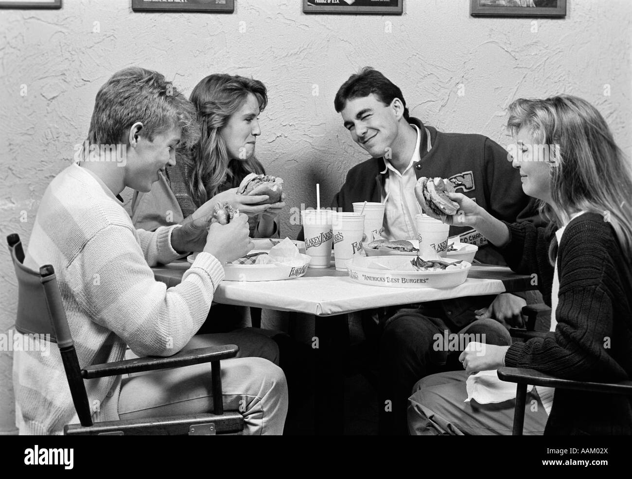1980s GROUP OF FOUR TEENS SITTING AROUND TABLE AT RESTAURANT CHATTING & EATING HAMBURGERS Stock Photo