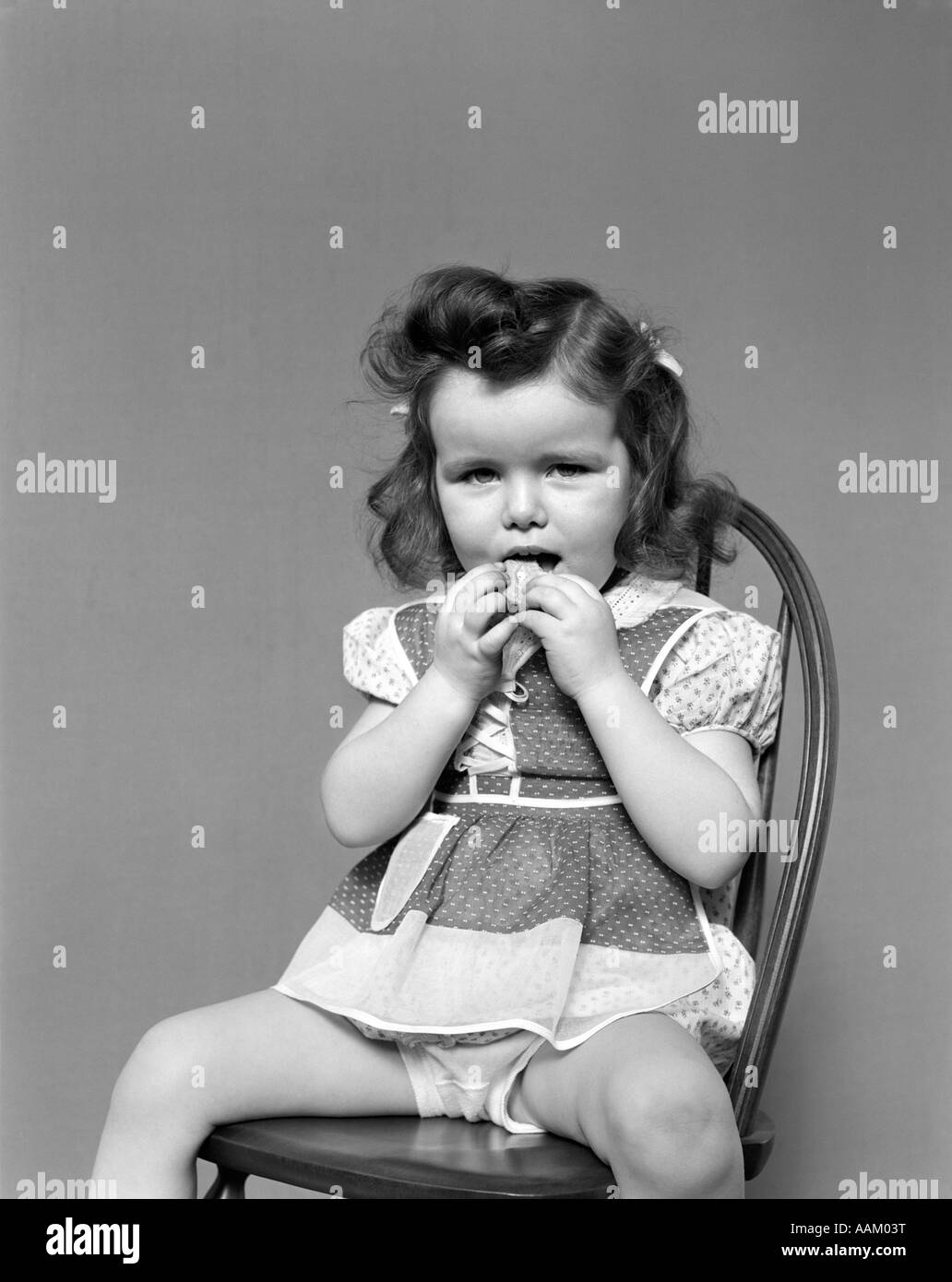 1930s TODDLER GIRL SIT IN WOODEN CHAIR POLKA DOT DRESS SHOWING UNDERPANTS EATING A COOKIE WITH BOTH HANDS VINTAGE Stock Photo