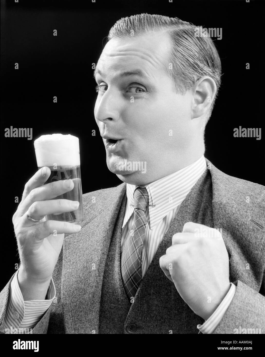 1930s MAN HOLDING FULL FOAMY GLASS OF BEER MAKING A FUNNY FACE WITH HIS THUMB HOOKED CONFIDENTLY UNDER HIS VEST LOOKING AT Stock Photo