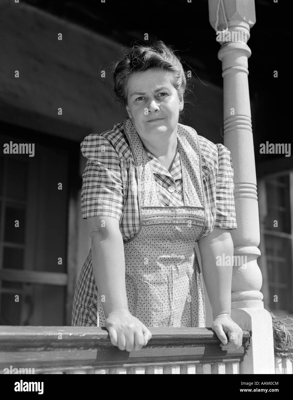 1940s OLDER WOMAN IN PLAID DRESS & APRON LEANING ON PORCH RAILING OF FARMHOUSE LOOKING AT CAMERA Stock Photo