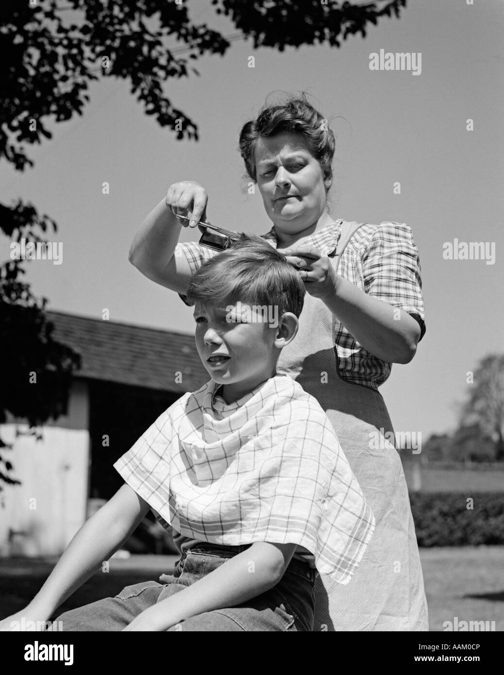 1940s FARMWIFE MOTHER CUTTING BOY SON'S HAIR OUTDOORS UNDER A TREE Stock Photo