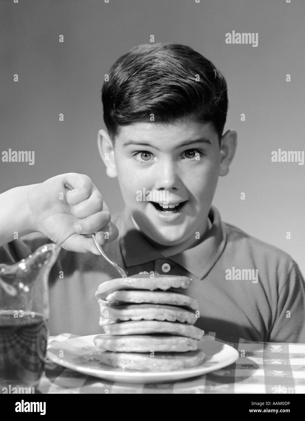 1960s SMILING PORTRAIT BOY SITTING EAGERLY BEHIND THICK STACK OF PANCAKES POKING FORK INTO TOP OF STACK LOOKING AT CAMERA Stock Photo