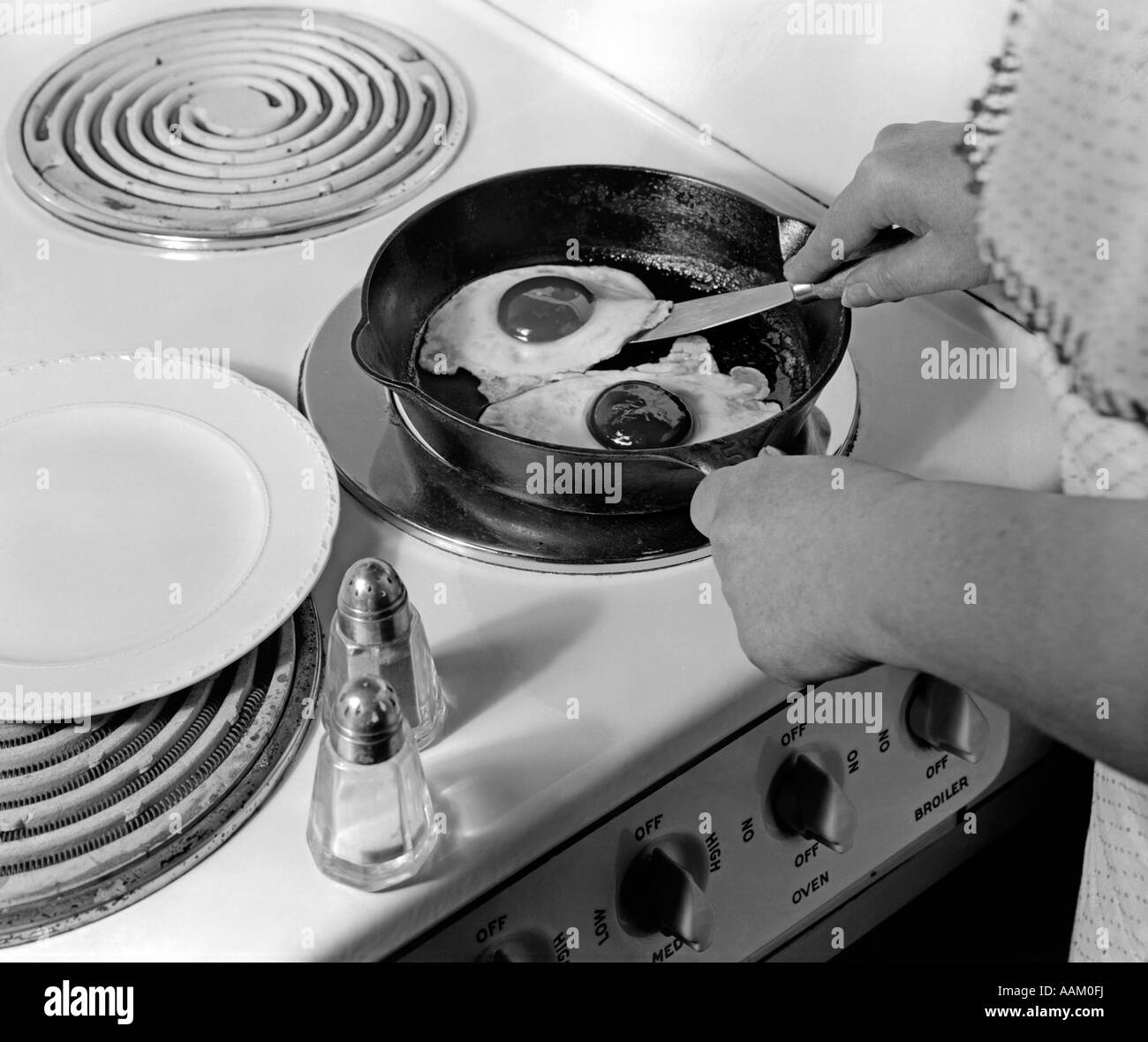 1940s 1950s WOMAN HANDS FRYING EGGS IN IRON SKILLET ON ELECTRIC STOVE SALT AND PEPPER SHAKERS Stock Photo