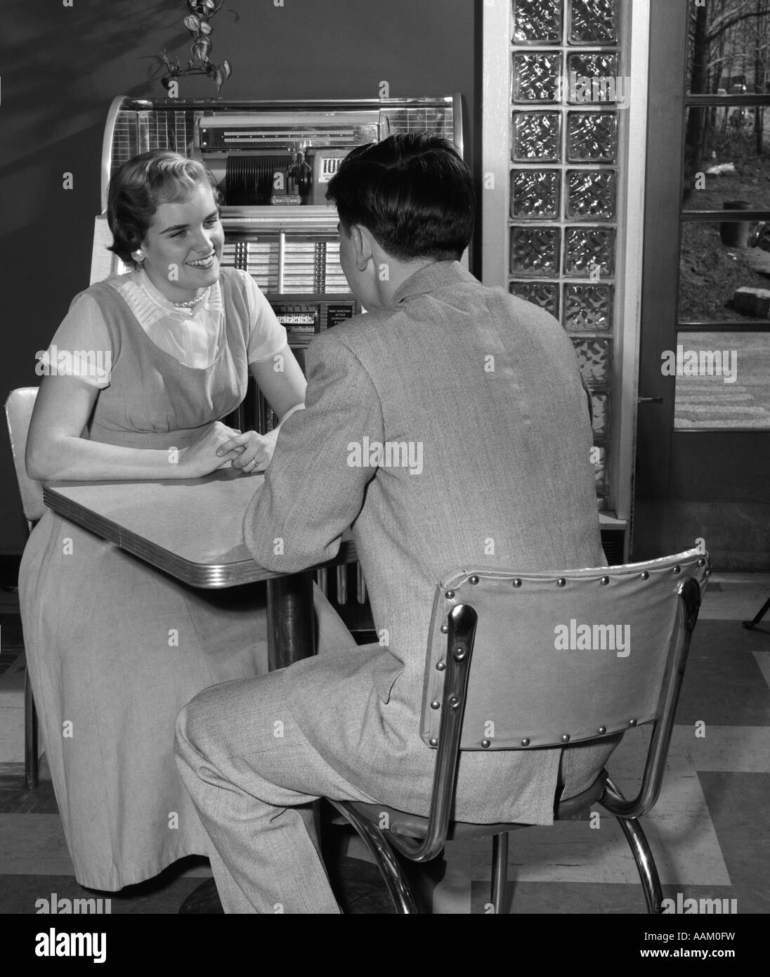 1950s TEEN COUPLE SITS AT TABLE IN DINER JUKEBOX IN BACKGROUND SMILING INDOOR Stock Photo