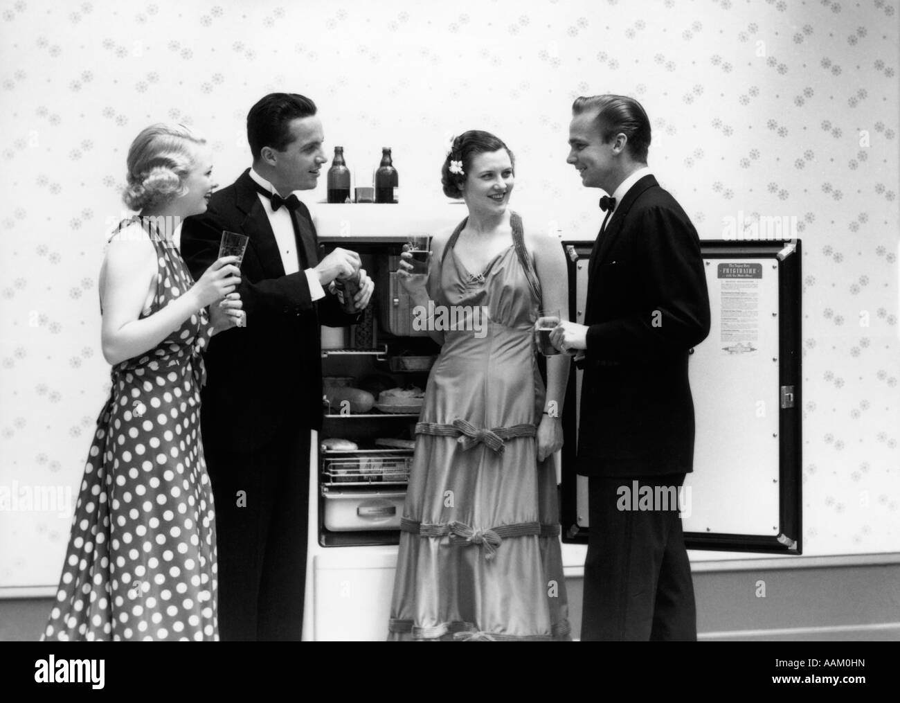 1930s 1940s TWO COUPLES FORMALLY DRESSED STANDING IN FRONT OF REFRIGERATOR WITH DOORS OPEN DRINKING BEER Stock Photo