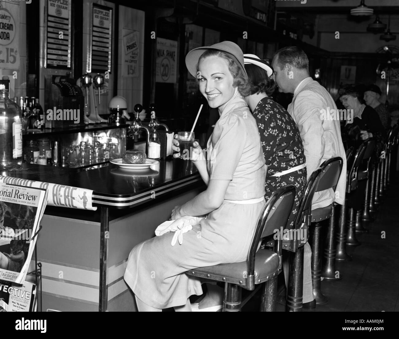 1940s SMILING WOMEN DRESSED IN LINEN DRESS HAT HOLDING GLASS COLA SITTING AT COUNTER SODA FOUNTAIN LOOKING AT CAMERA Stock Photo
