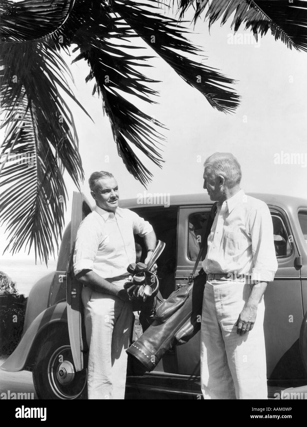 1930s 2 ELDERLY MEN STANDING BY CAR CARRYING GOLF BAG PALM TREE FROND IN CORNER Stock Photo