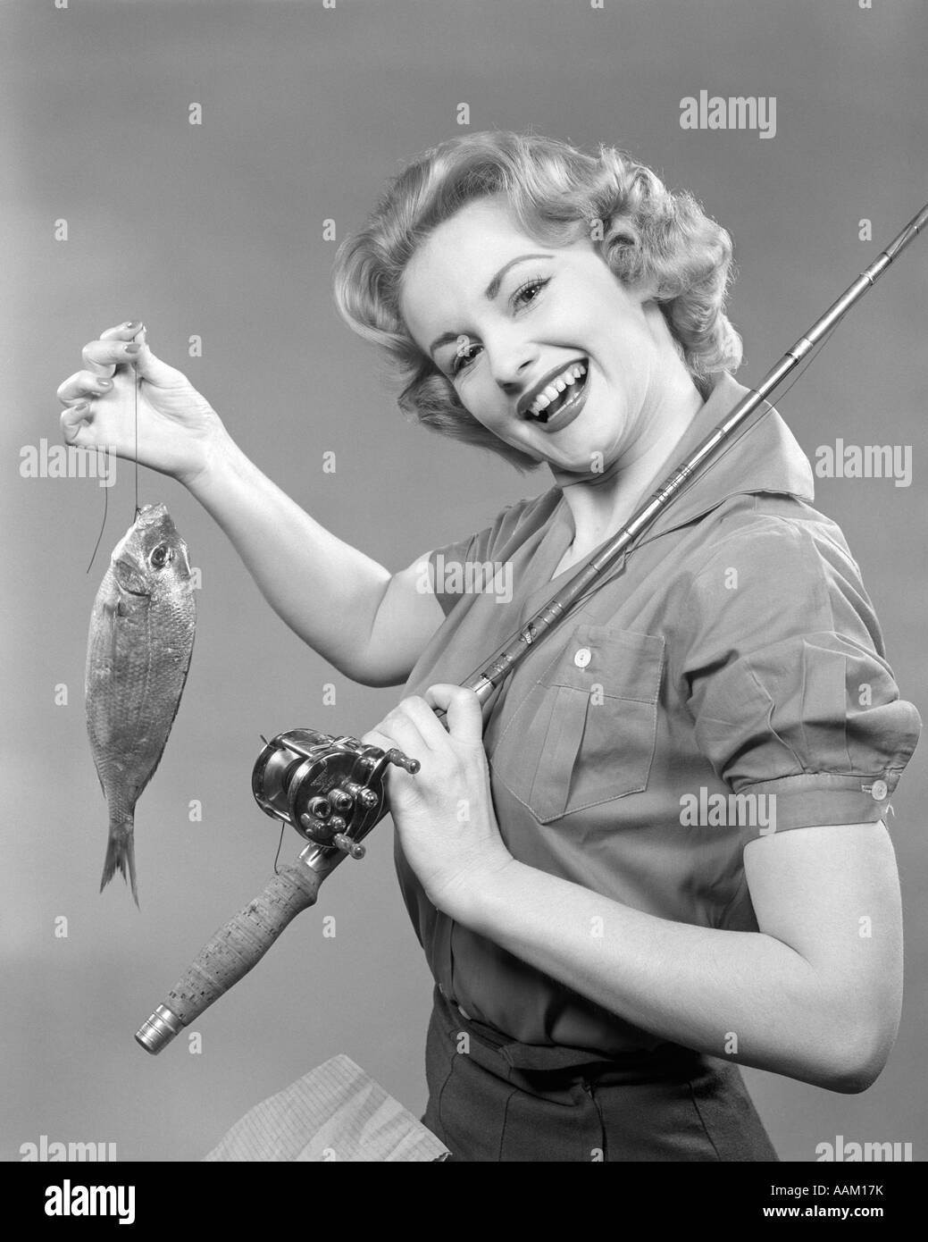 1950s SMILING WOMAN WITH A FISHING ROD OVER HER SHOULDER HOLDING UP A FISH LOOKING AT CAMERA Stock Photo