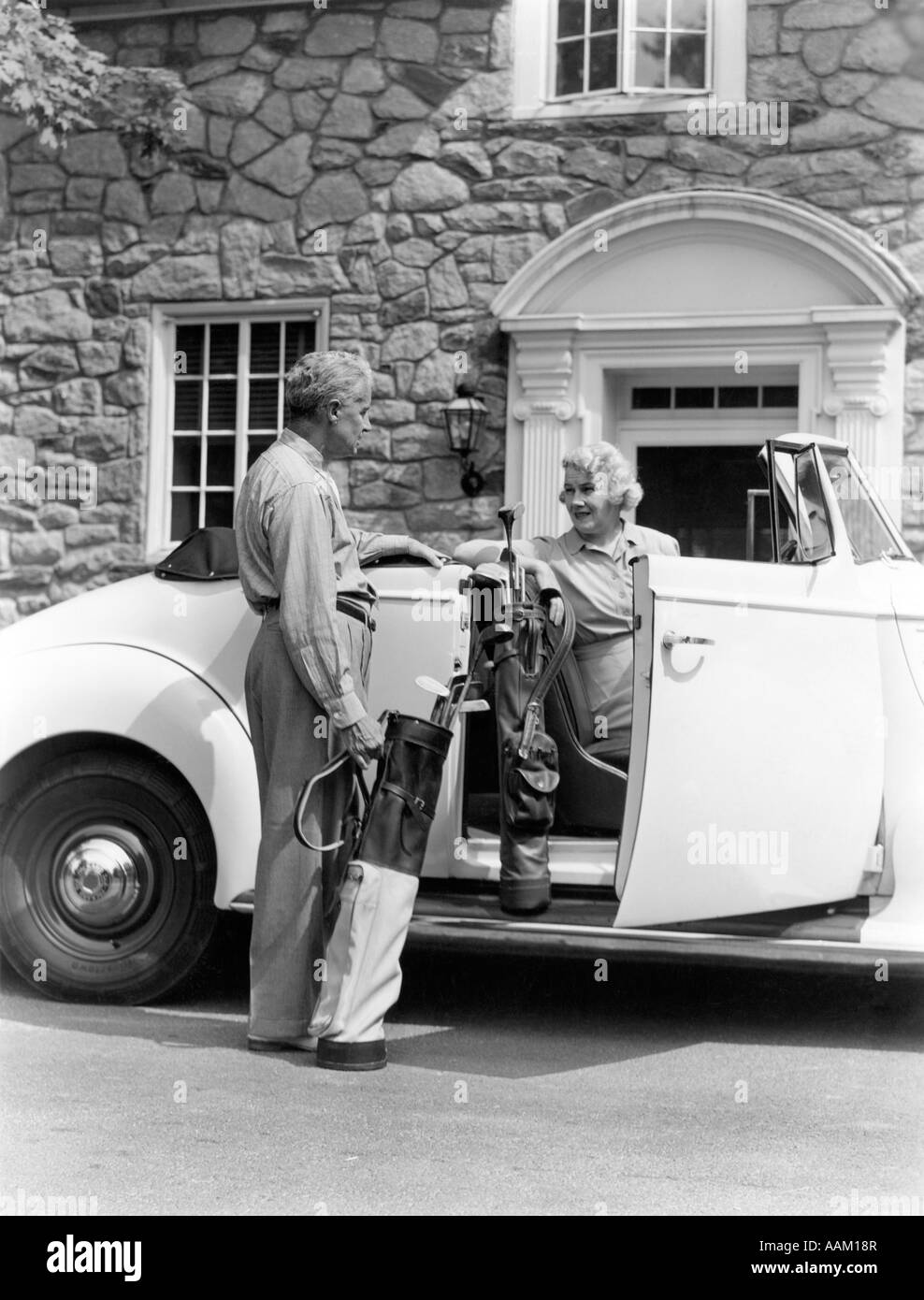 1940s SENIOR RETIRED COUPLE LOADING GOLF CLUBS INTO WHITE CONVERTIBLE CAR IN FRONT STONE HOUSE Stock Photo