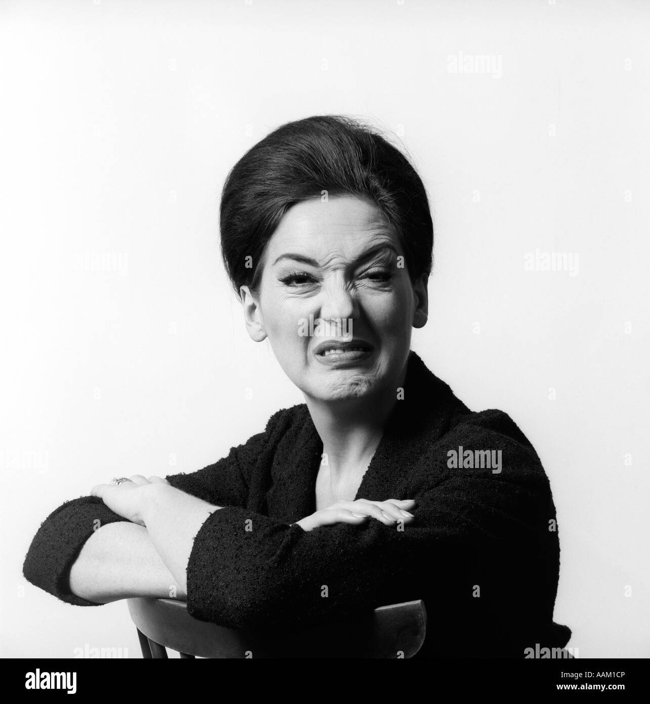 1960s 1970s BRUNETTE MIDDLE AGED WOMAN MAKING FUNNY FACE LOOKING AT CAMERA WITH WRINKLED NOSE AND ARMS FOLDED ON BACK OF CHAIR Stock Photo