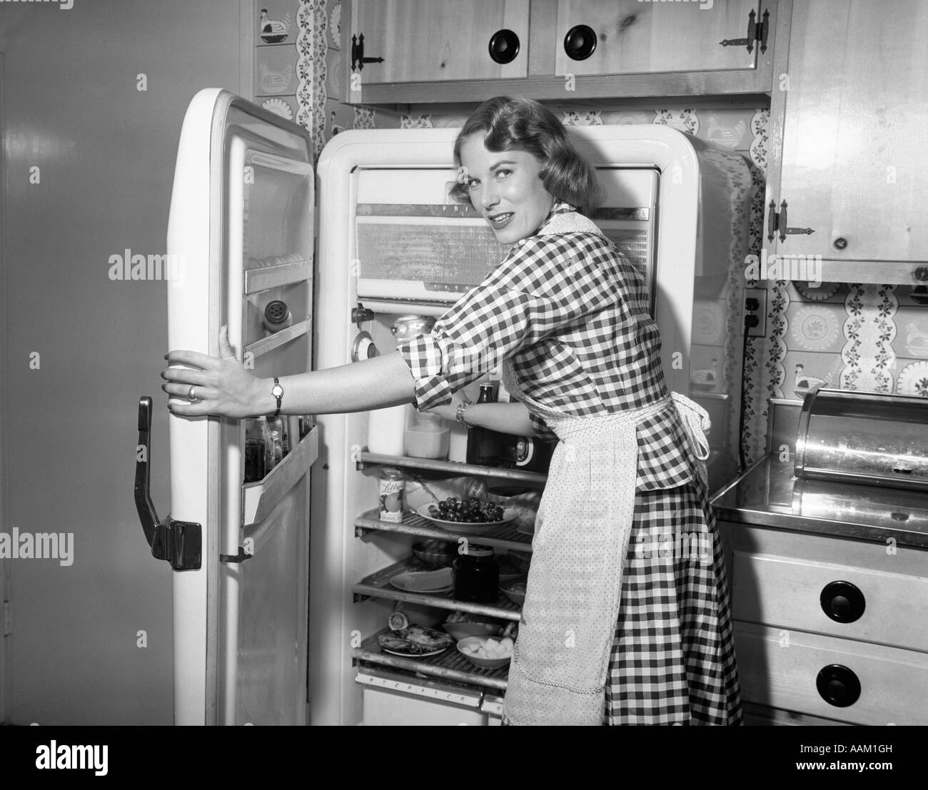 1950s WOMAN WEARING GINGHAM DRESS AND APRON OPENING REFRIGERATOR DOOR Stock Photo