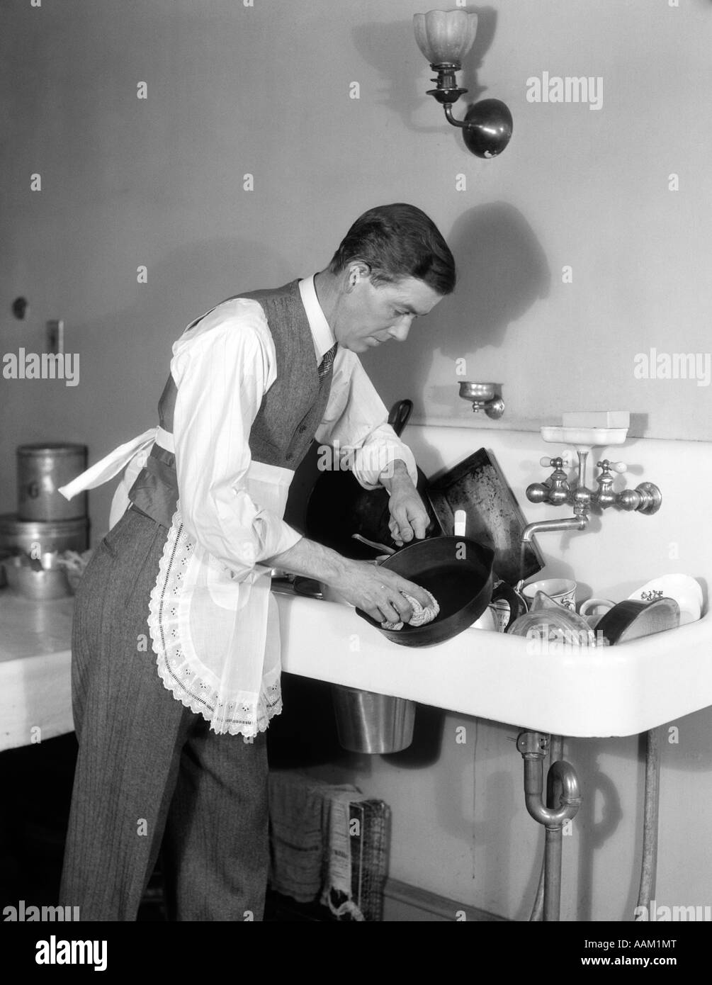 1930s MAN DRESSED IN SHIRT TIE VEST & APRON BENT OVER A SINK FULL OF DISHES CLEANING AN IRON SKILLET WITH A DISHCLOTH Stock Photo