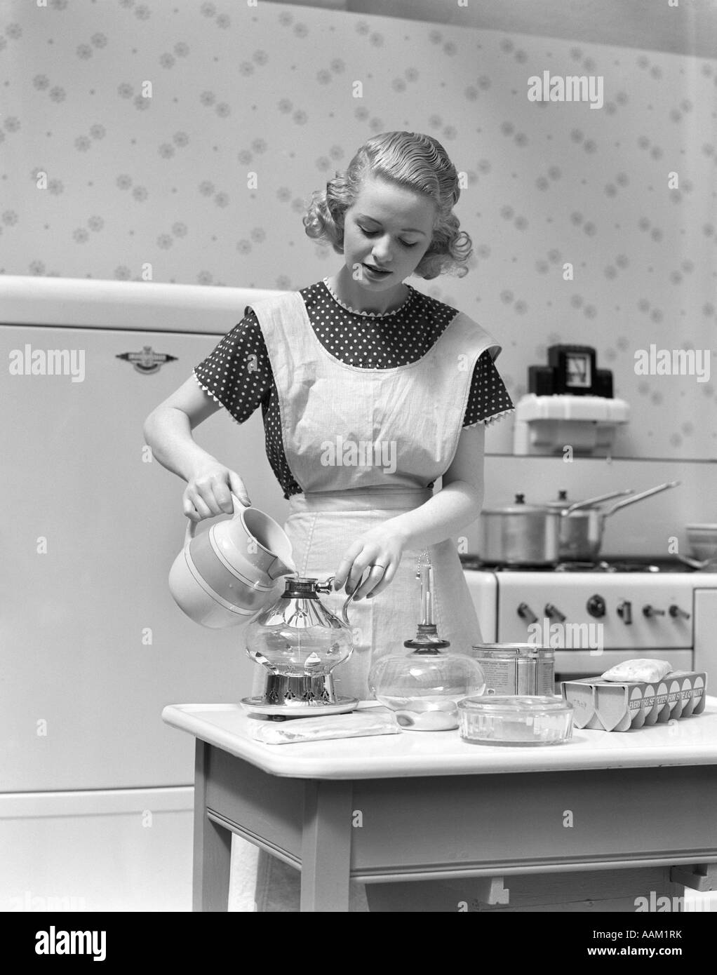 1930s WOMAN IN KITCHEN WEARING APRON MAKING BREAKFAST POURING WATER INTO COFFEE POT Stock Photo