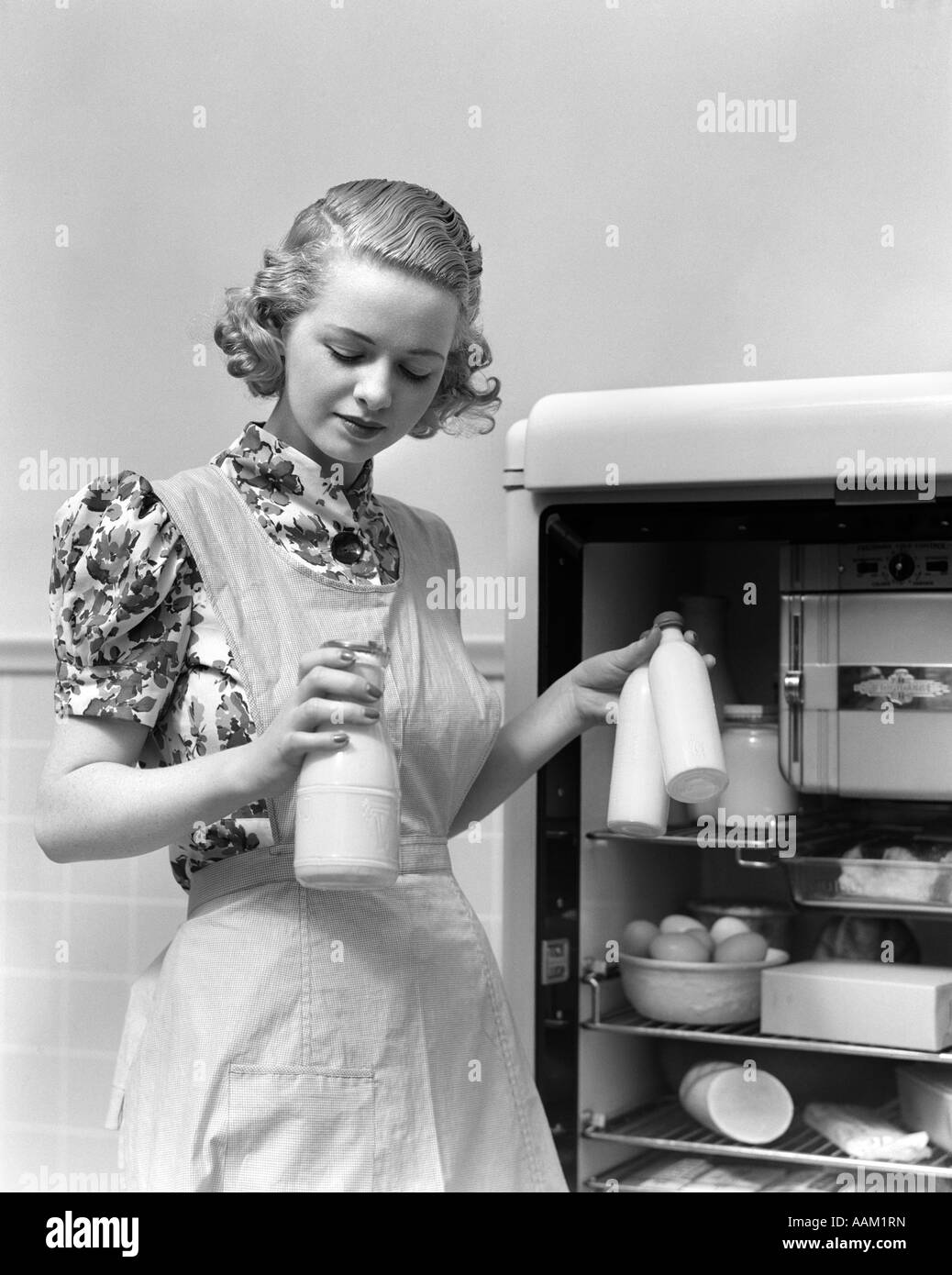 1930s WOMAN WEARING A WHITE APRON TAKING MILK OUT OF THE REFRIGERATOR Stock Photo