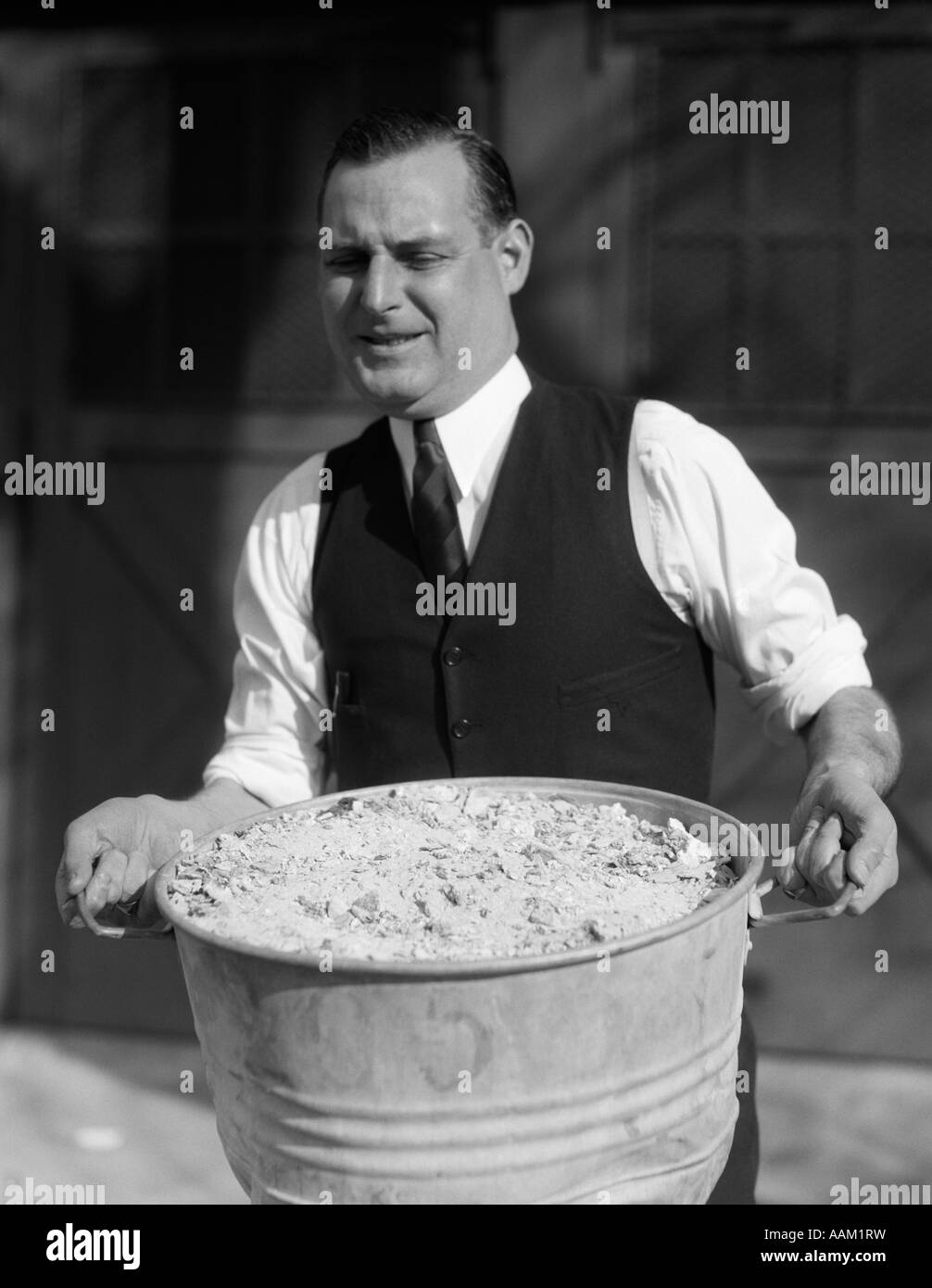 1930s MAN IN SHIRT VEST & TIE STRAINING AS HE CARRIES A HEAVY ASH CAN FULL OF ASHES Stock Photo