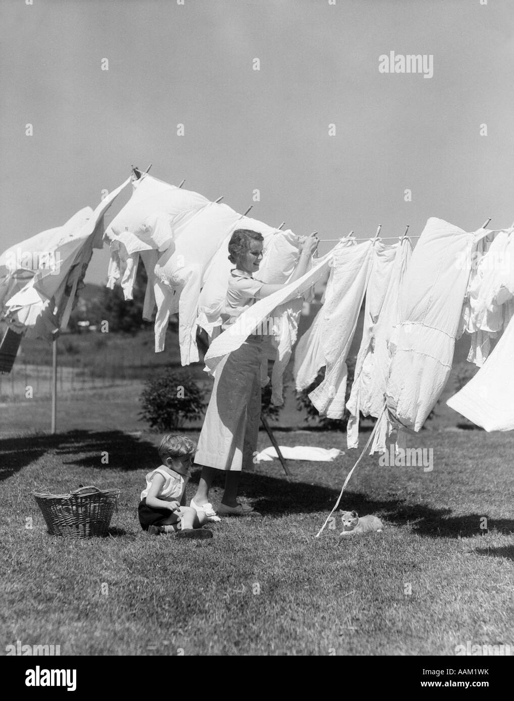 1930s WOMAN IN WHITE COTTON DRESS HANGING A FULL LINE OF CLOTHES IN THE BREEZE WITH A LITTLE BOY & CAT AT HER FEET Stock Photo