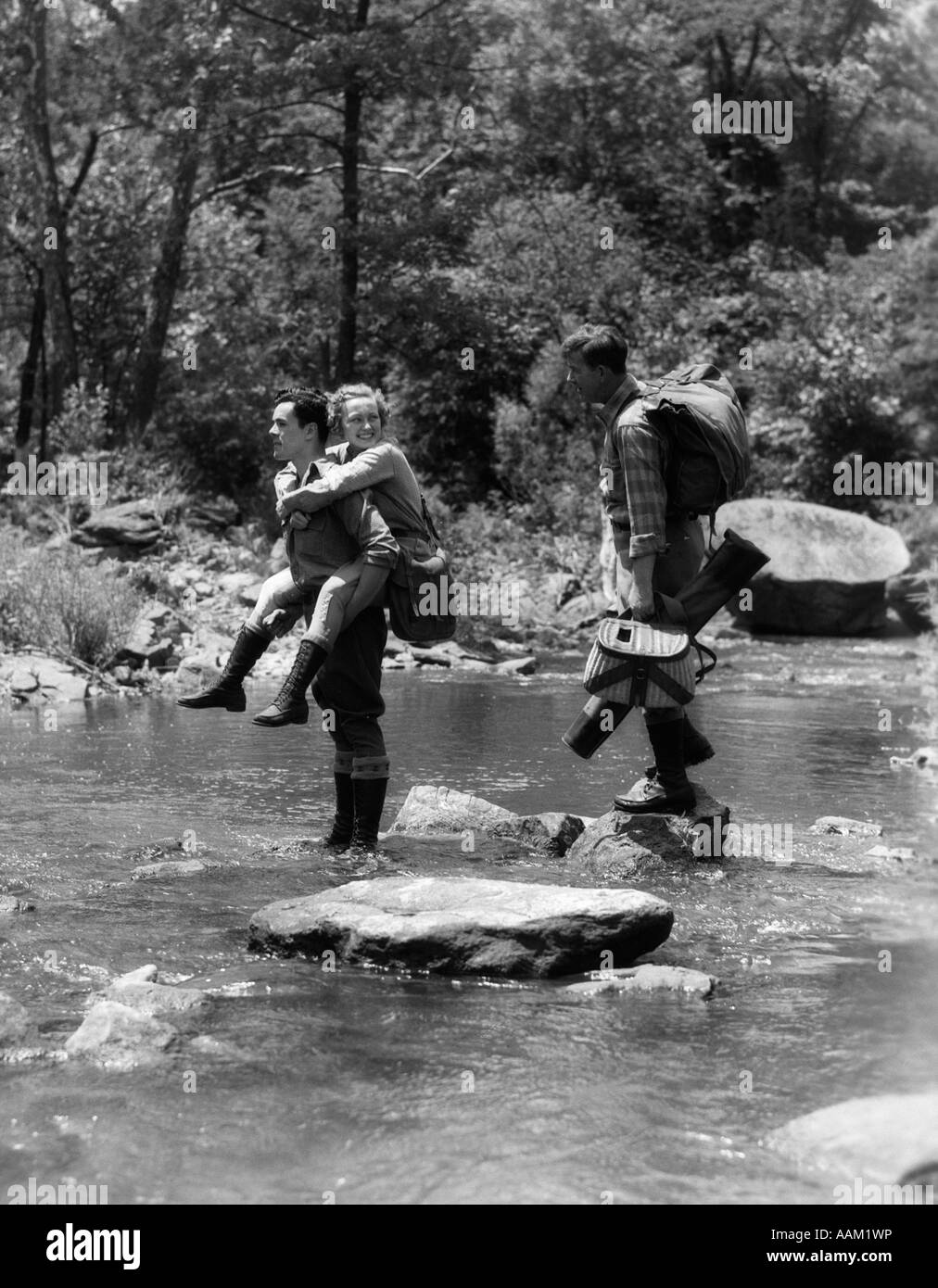 1930s 3 PEOPLE CROSSING FORDING STREAM ONE MAN CARRIES WOMAN PIGGY BACK OTHER CARRIES ANGLING FISHING GEAR STEPPING STONES WATER Stock Photo