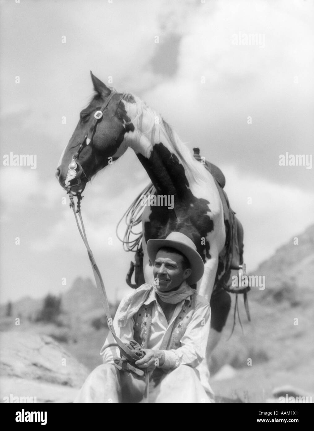 1930s COWBOY SITTING IN FRONT OF HORSE HOLDING REINS SPOTTED PAINT PINTO Stock Photo