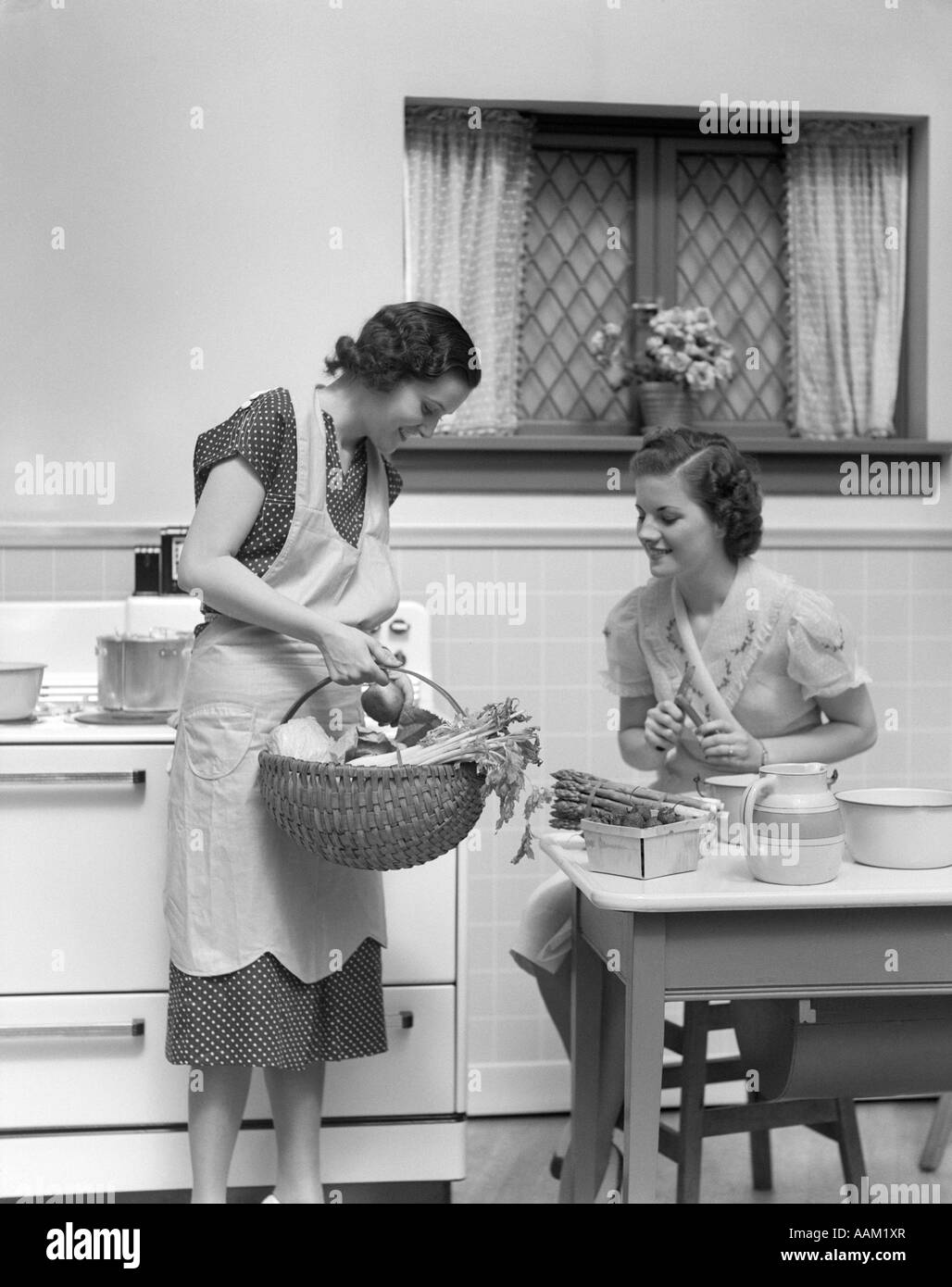 1930s 1940s TWO WOMEN AT KITCHEN TABLE WITH A BASKET OF VEGETABLES ASPARAGUS STOVE Stock Photo