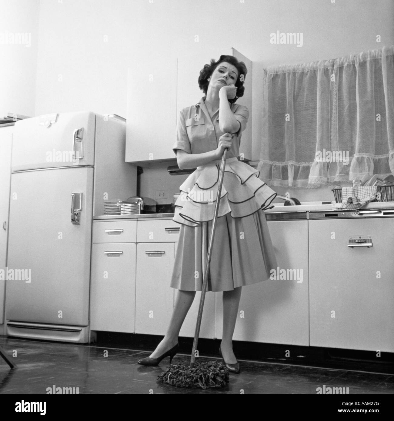 1950s BORED WOMAN HOUSEWIFE WEARING APRON LEANING ON MOP ON KITCHEN FLOOR Stock Photo