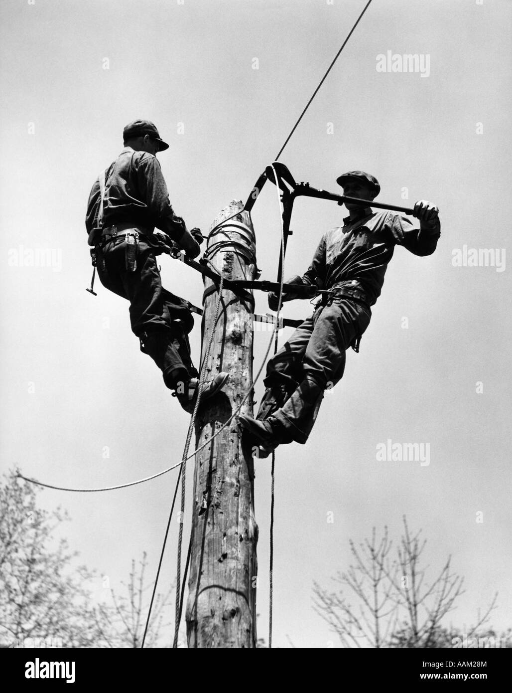 1930s 1940s TWO MEN WORKING ON ELECTRICAL POWER POLE CUTTING WIRE Stock
