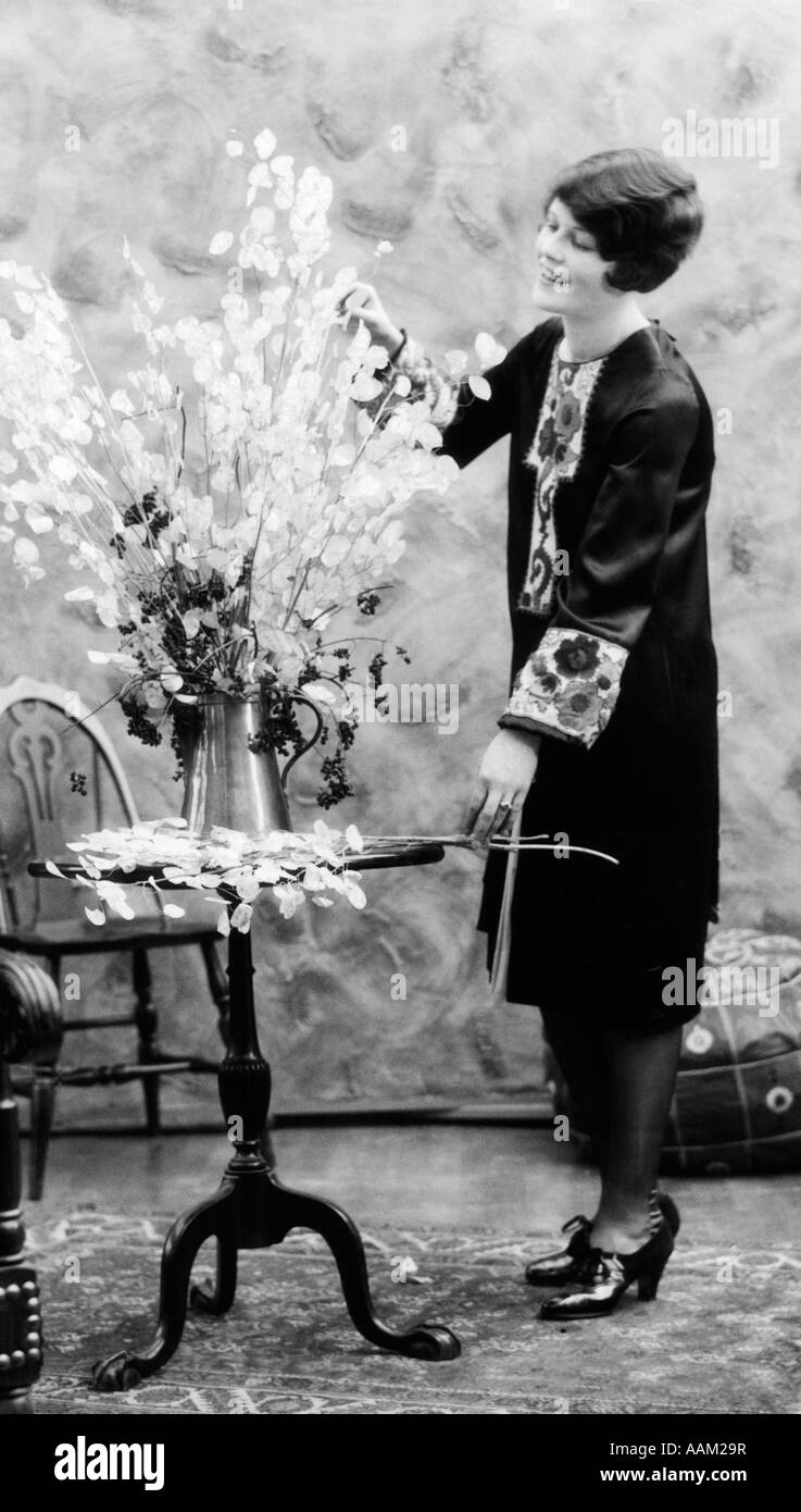 1920s WOMAN MAKING FLOWER ARRANGEMENT STANDING BY CHIPPENDALE TABLE ON ORIENTAL CARPET Stock Photo