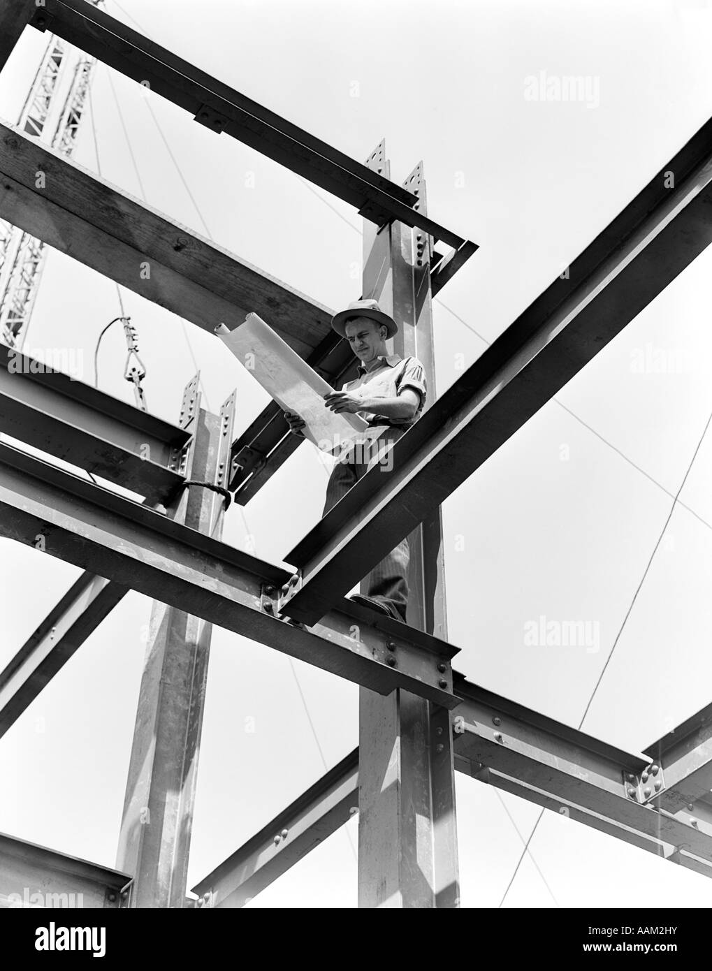 1940s MAN IN DRESS HAT & WORK SHIRT HOLDING BLUE PRINT WITH BOTH HANDS WHILE STANDING ON STEEL FRAMEWORK OF A LARGE BUILDING Stock Photo