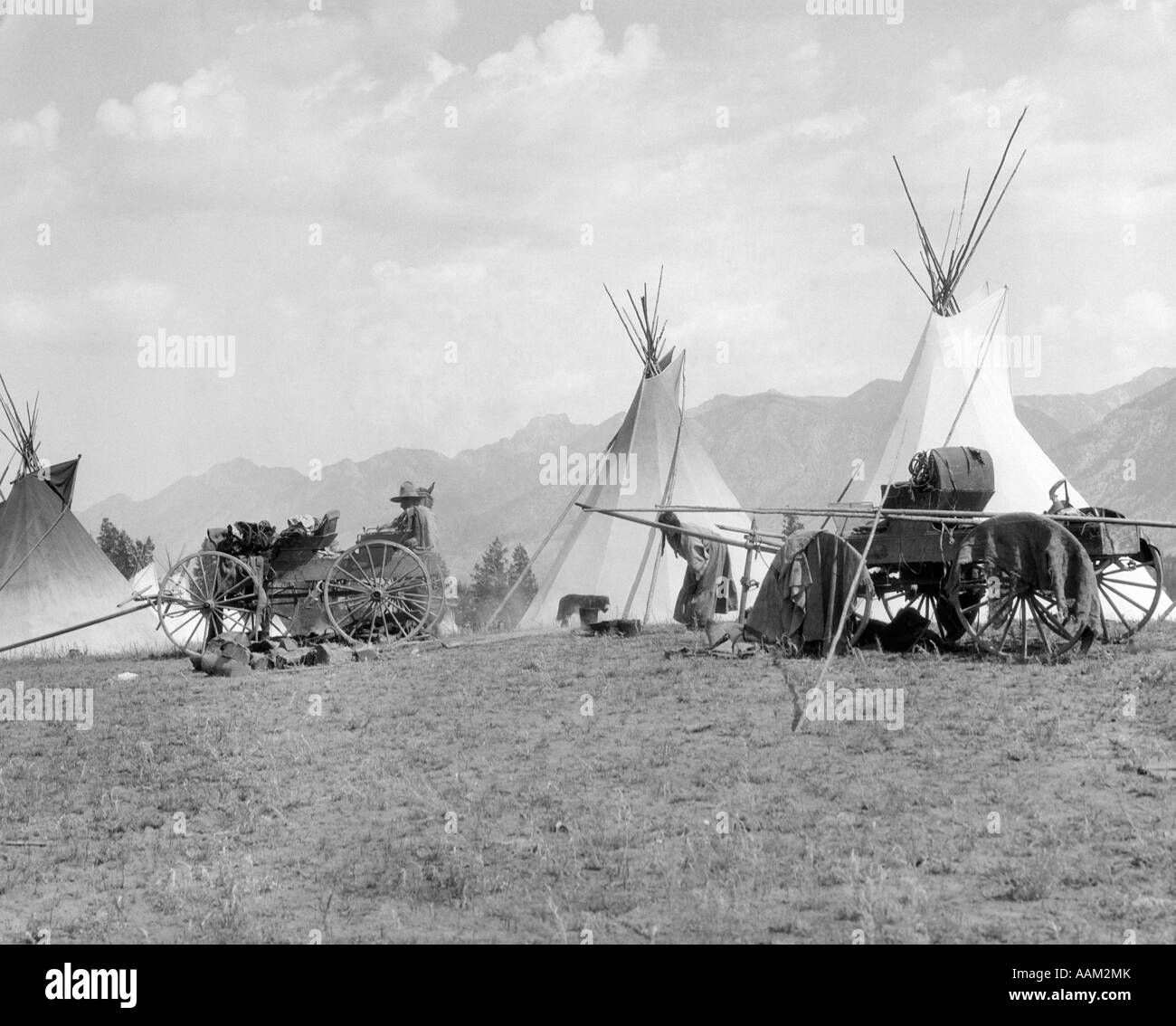 1920s 1930s NATIVE AMERICAN KOOTENAI NATIVE AMERICAN INDIAN CAMP VILLAGE TIPI TEEPEE WAGONS MOUNTAINS IN BACKGROUND BRITISH Stock Photo