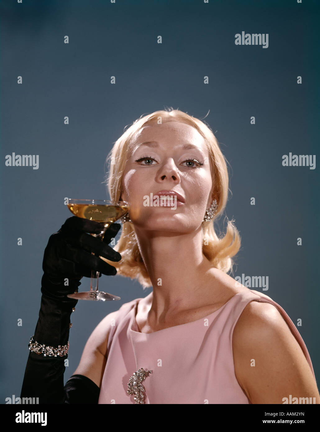 1960s FASHION ELEGANT BLOND WOMAN BLACK OPERA GLOVES BRACELET JEWELRY EARRINGS BROOCH HOLD GLASS OF CHAMPAGNE Stock Photo