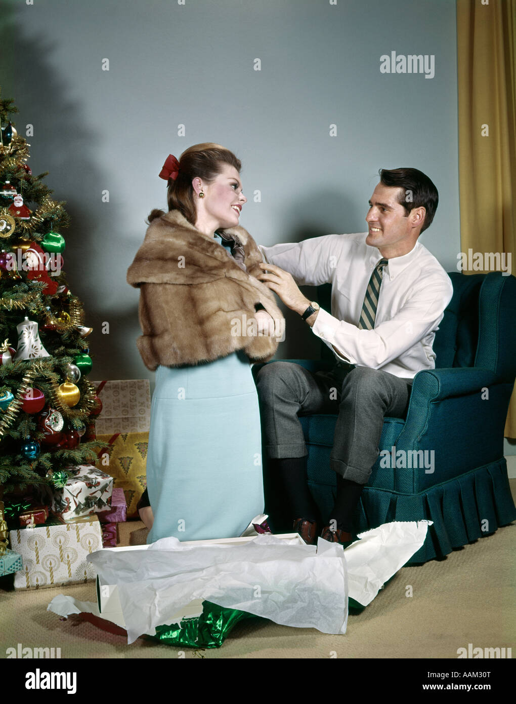 1970s LIFESTYLE COUPLE MAN ADJUSTING NEW HOLIDAY GIFT FUR JACKET ON WIFE IN LIVING ROOM BESIDE CHRISTMAS TREE Stock Photo