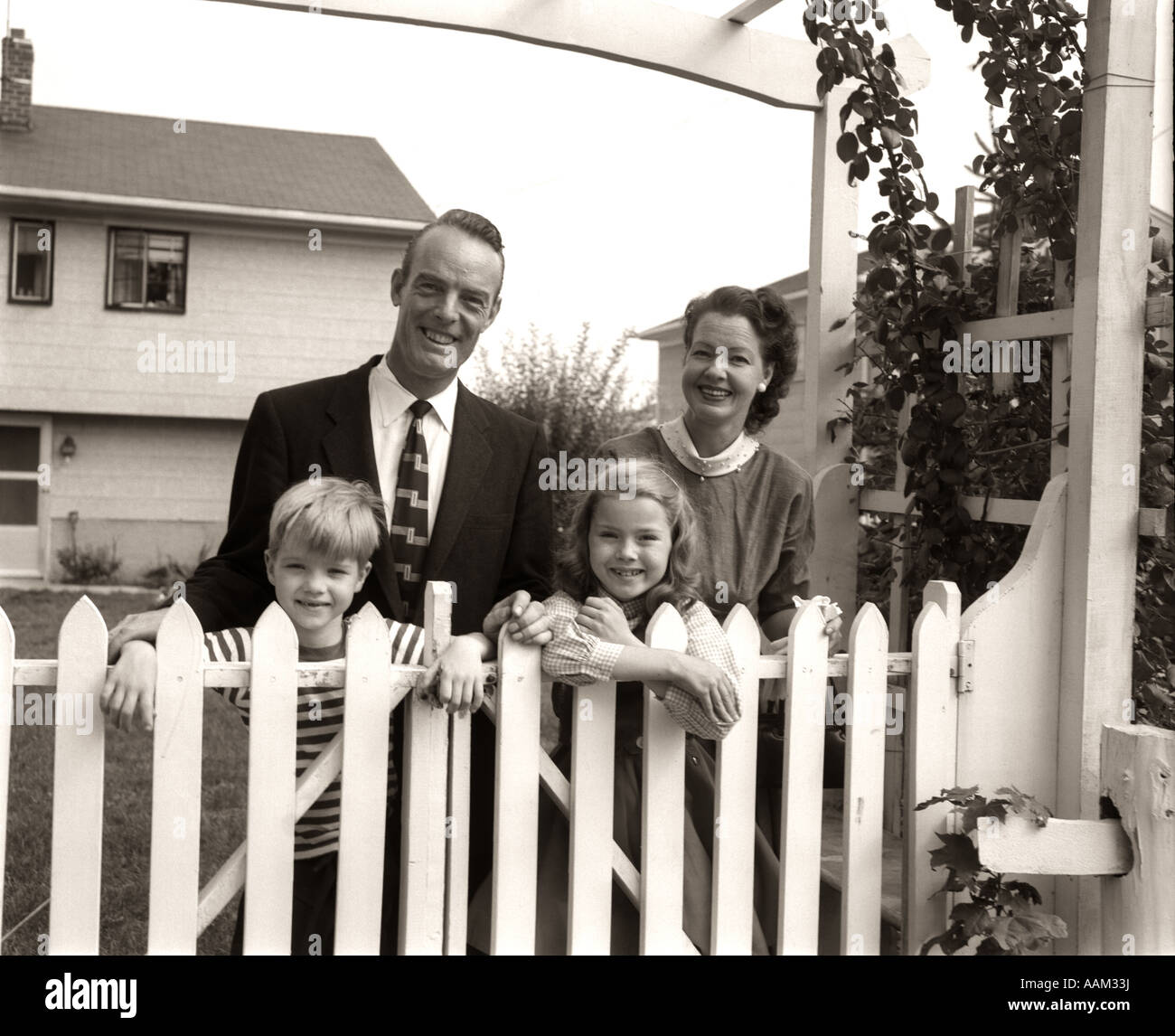 1950s FAMILY OF FOUR BEHIND PICKET FENCE IN BACKYARD SMILING AT CAMERA Stock Photo
