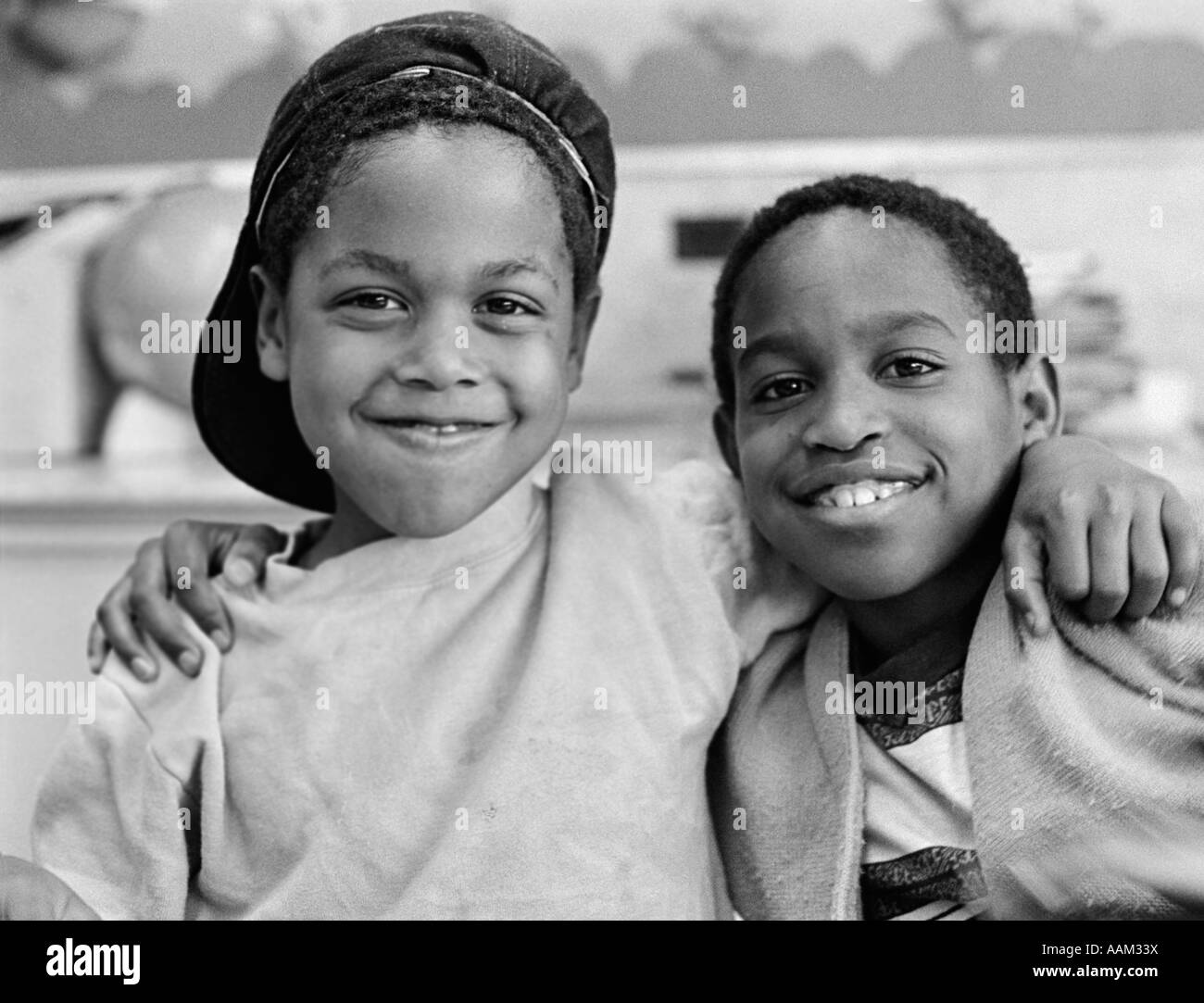 1980s TWO AFRICAN AMERICAN BOYS SMILING WHILE EMBRACING SHOULDER TO SHOULDER ONE BOY HAS CAP ON BACKWARDS OUTSIDE Stock Photo