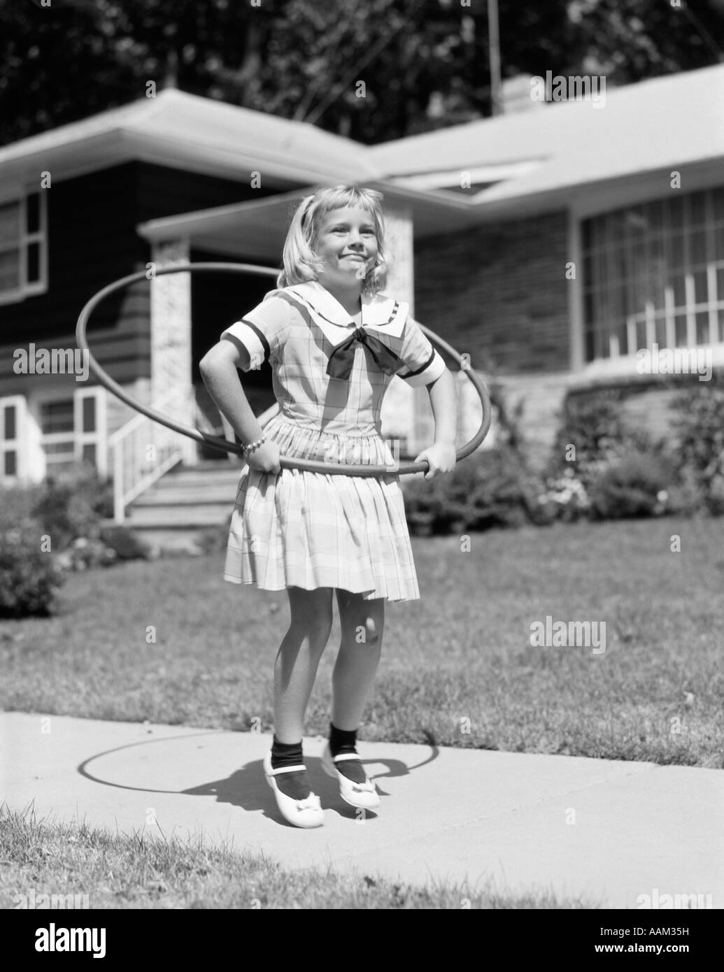 1950s GIRL IN DRESS ON SUBURBAN SIDEWALK USING THE HULA HOOP Stock Photo