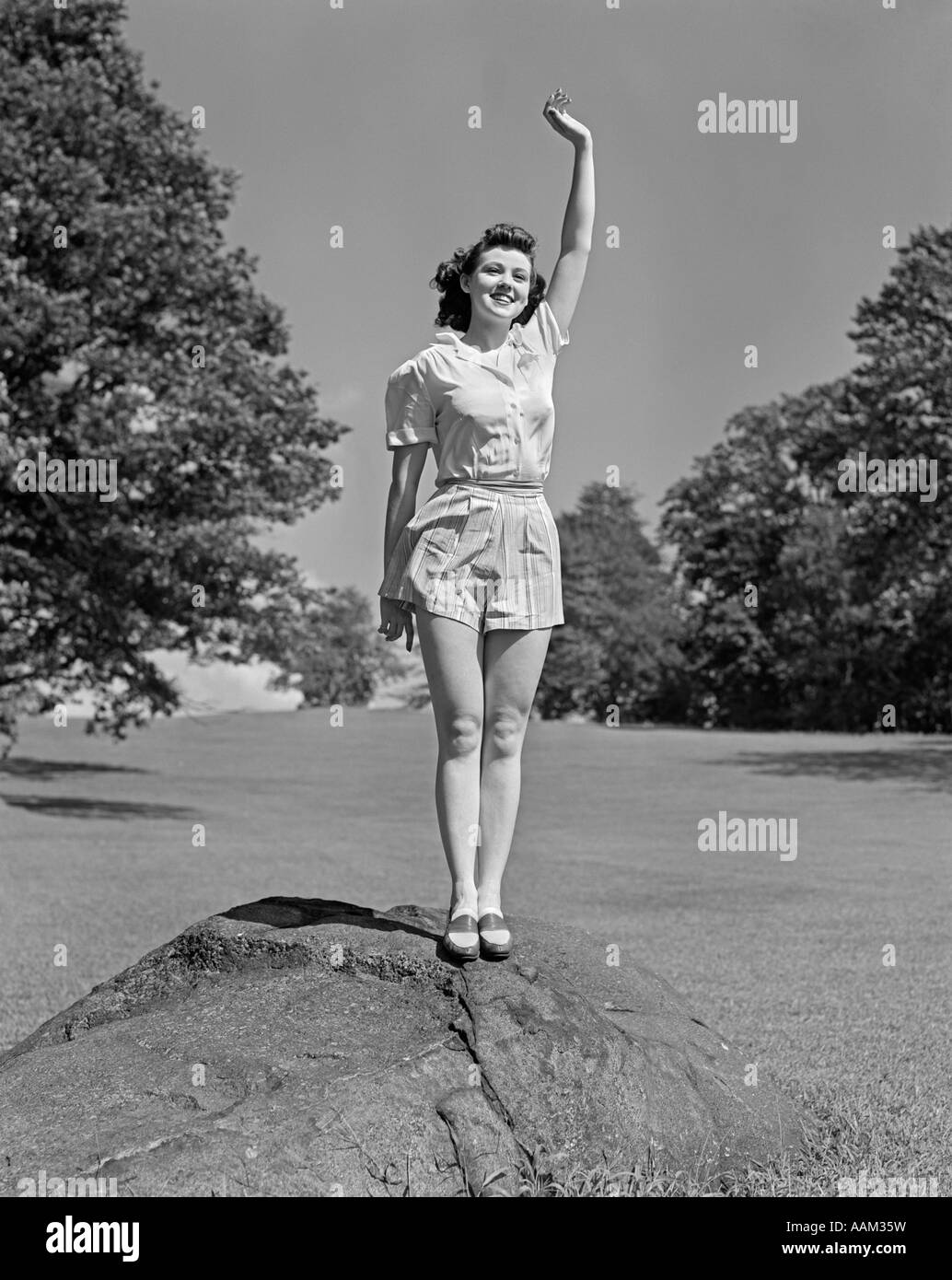 1940s SMILING TEEN WOMAN WEARING SHORTS STANDING ON ROCK WAVING HER HAND ABOVE HER HEAD Stock Photo