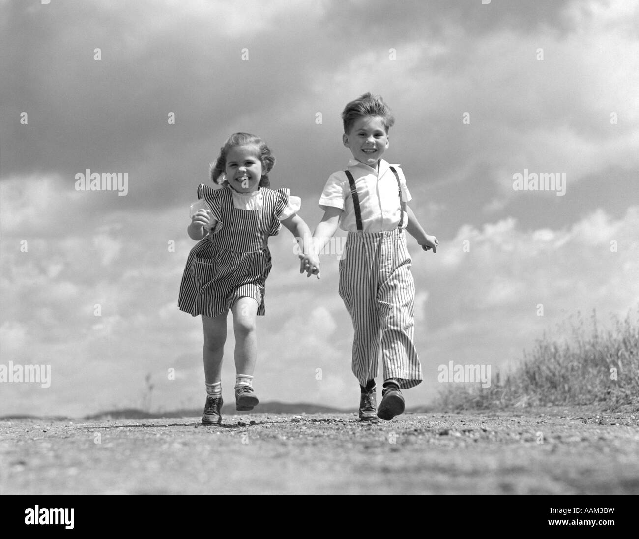 1940s 1950s BOY GIRL HOLDING HANDS RUNNING DOWN DIRT ROAD CLOUDS SKY BEHIND HAPPY SMILING COUPLE FRIENDS FRIENDSHIP ROMANCE Stock Photo
