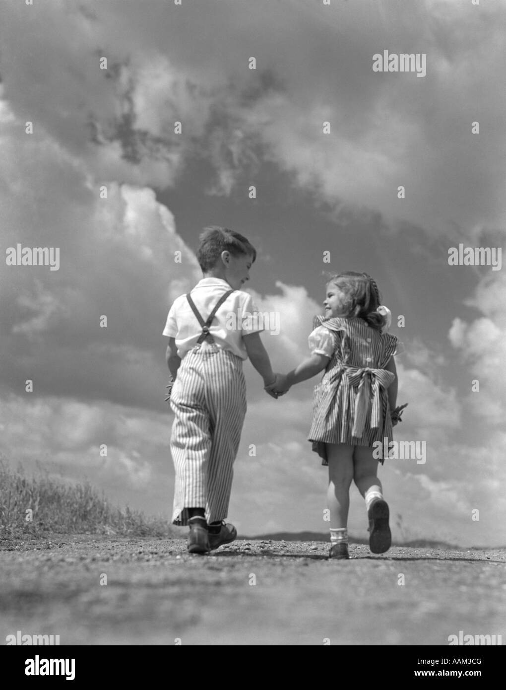 1940s BOY GIRL HOLDING HANDS WALKING UP HILL INTO BRIGHT SKY FULL OF CLOUDS Stock Photo