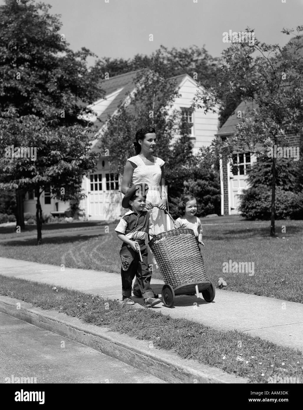 1950s MOM ON SIDEWALK PUSHING WICKER SHOPPING BASKET WITH DAUGHTER AND SON IN COWBOY HAT WALKING BESIDE HER Stock Photo