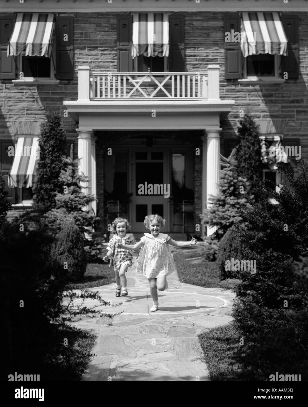 1930s 2 GIRLS IN SUMMER DRESSES RUNNING TO CAMERA ON FLAGSTONE SIDEWALK IN FRONT OF HOUSE Stock Photo